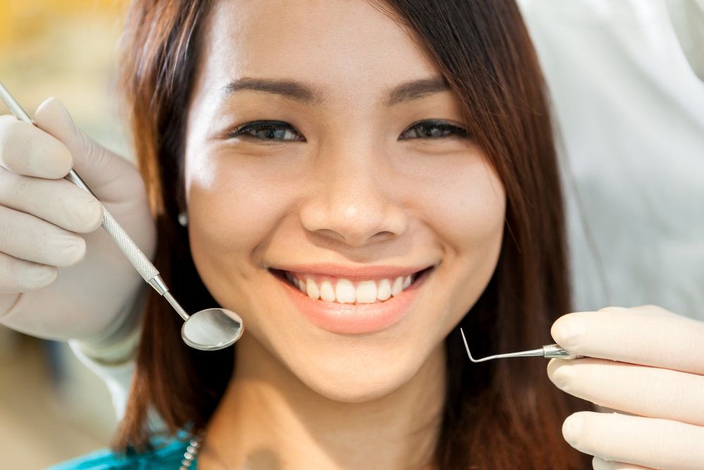 <p>A closeup of a young woman&#39;s face with a dentist holding a mirror and dental pick in front of her.</p>
